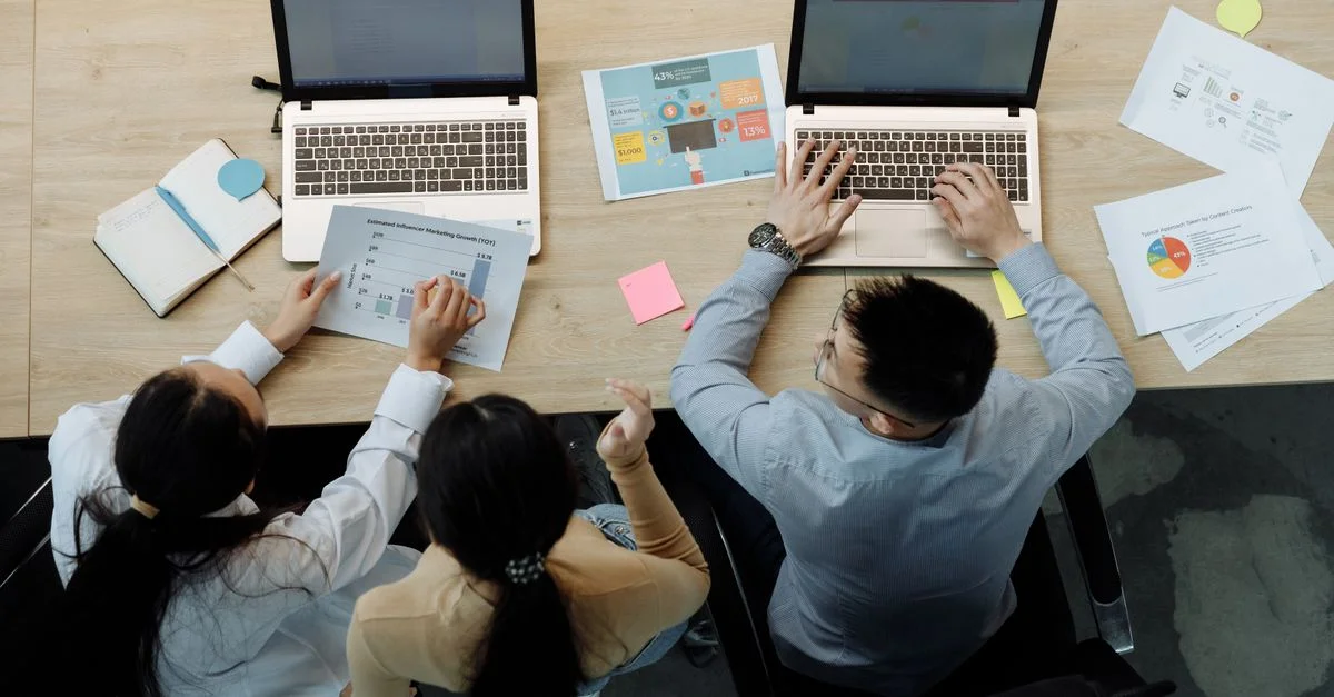 Trois collègues travaillant sur des ordinateurs portables et des documents à un bureau, collaborant sur des projets d'affaires.
