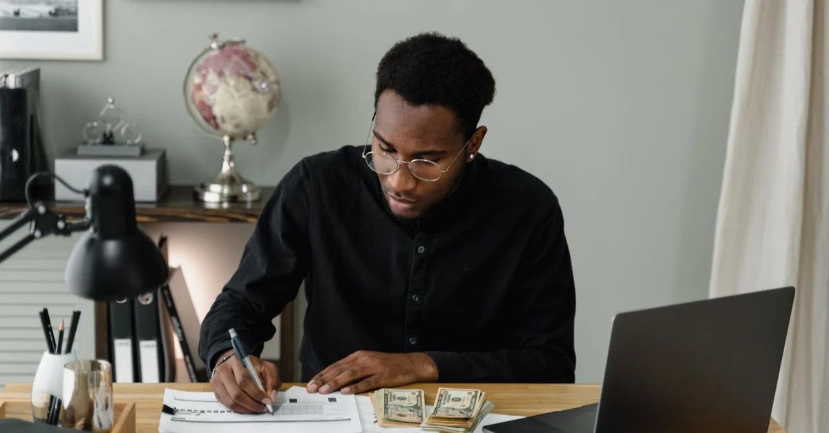 Un homme adulte calcule des dépenses, utilisant un ordinateur portable et des documents à un bureau dans un cadre de bureau à domicile.